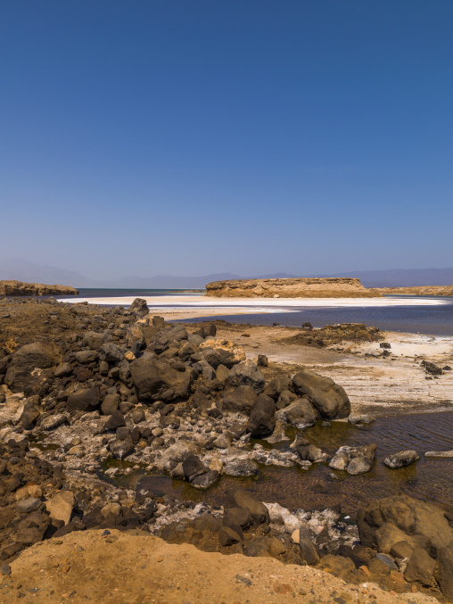 Salt-encrusted volcanic rocks line the shore of a turquoise crater lake, Dikhil Region, Lake Assal, Djibouti