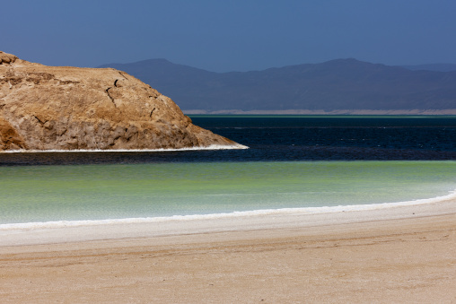 A rocky point stretches into the green water of Lake Assal beyond a sandy beach, Dikhil Region, Lake Assal, Djibouti