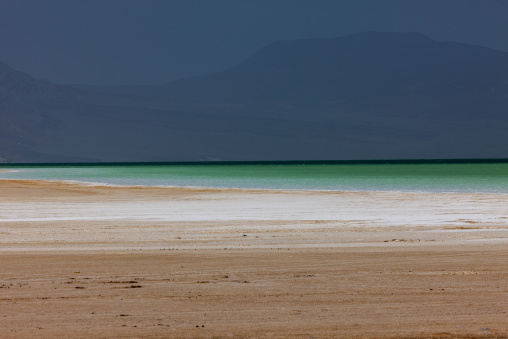 White salt flat stretches toward the green waters of Lake Assal beneath distant mountains, Dikhil Region, Lake Assal, Djibouti