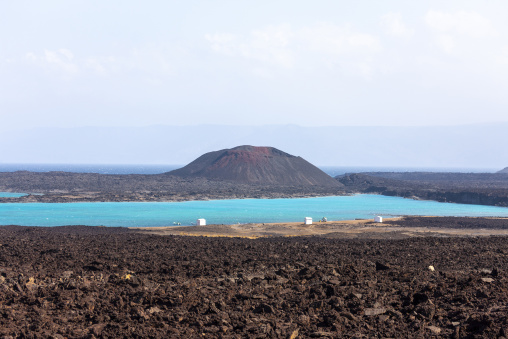 A red-tinged volcanic cone overlooks a turquoise lagoon bordered by black lava fields, Tadjourah Region, Goubet Al-kharab, Djibouti