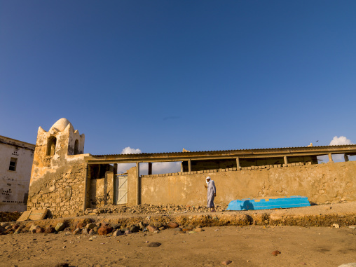 A man in traditional dress walks past crumbling coastal ruins with a blue fishing boat on the shore, Tadjourah Region, Tadjoura, Djibouti