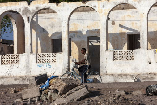 A man rides a bicycle past the weathered arches of an old ruined building as a goat wanders nearby, Tadjourah Region, Tadjoura, Djibouti