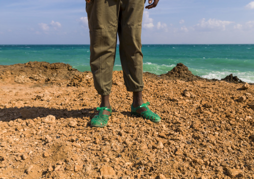 A person wearing green jelly sandals stands on a rocky coastline, Obock Region, Khor angar, Djibouti