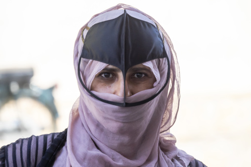 Extreme close-up front-facing portrait of an Omani woman wearing a pale pink headscarf and black leather face mask against a bright background, Ad Dakhiliyah Region, Nizwa, Oman