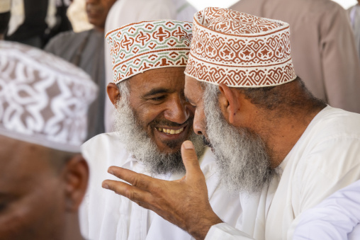 Close-up of two elderly Omani men wearing embroidered kuma caps laughing and gesturing together at the Nizwa market, Ad Dakhiliyah Region, Nizwa, Oman