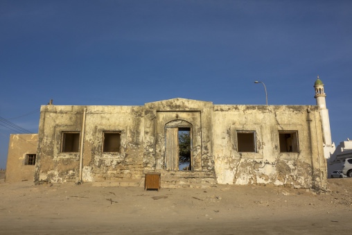 Abandoned crumbling house with broken windows and an ajar wooden door beneath a distant mosque minaret, Dhofar Governorate, Mirbat, Oman