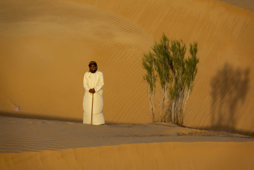 Close-up portrait of a man with a wrapped headscarf and cream robe standing near a green desert shrub, Dhofar Governorate, Rub al Khali, Oman