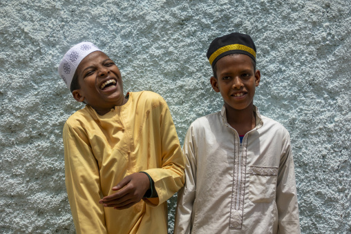 Two young Ethiopian boys wearing traditional kufi caps laugh together against a textured wall, Harari Region, Harar, Ethiopia