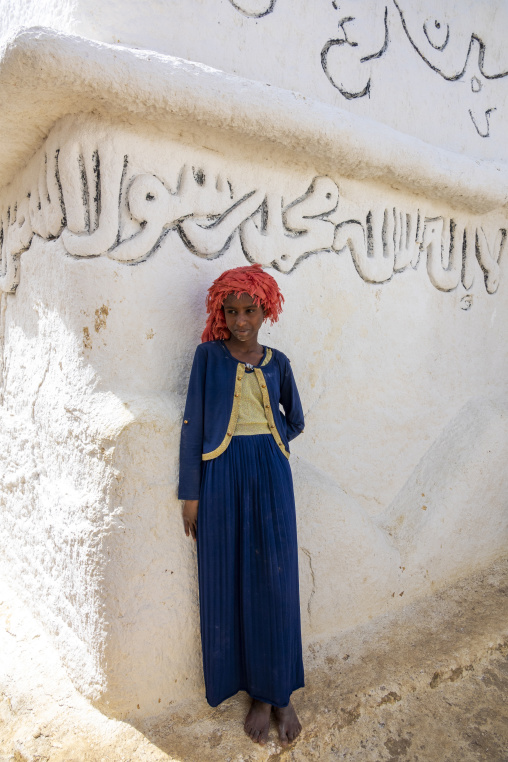 A young Sufi pilgrim in a red headscarf stands beside a wall inscribed with Islamic calligraphy, Oromia, Sheik Hussein, Ethiopia