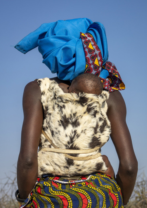 Rear view of a Mucubal woman carrying her baby wrapped in animal fur on her back while wearing a blue headscarf, Namibe Province, Virei, Angola