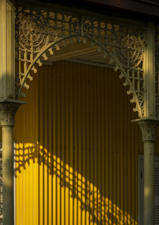Shadow of a tree branch falls across the yellow wall beneath an arched ironwork trellis of the Palacio de Ferro, Luanda Province, Luanda, Angola
