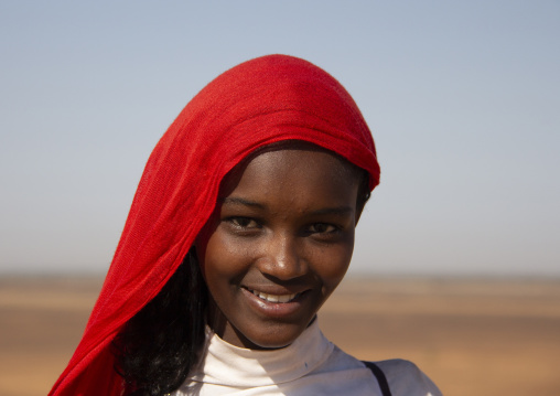 A smiling young woman wears a red headscarf in front of a desert landscape, Northern State, Meroe, Sudan
