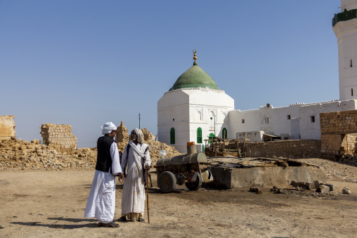 Two men stand near a donkey cart carrying water barrels in front of a domed mosque in the old town, Red Sea State, Suakin, Sudan
