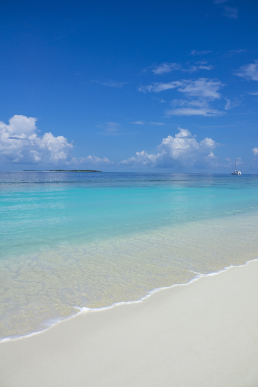 A distant sailboat drifts near a small island beyond a turquoise lagoon and white sand beach, Baa Atoll, Soneva Fushi, Maldives