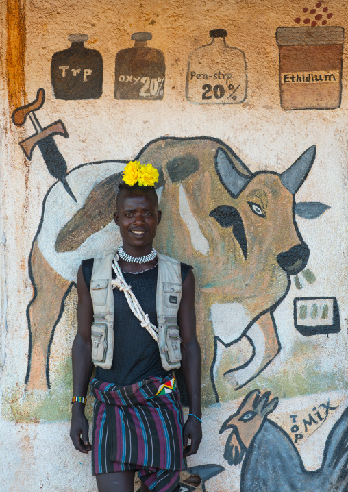 Smiling bana tribe man with yellow plastic flower in the hair in front of a mural depicting a cow, Omo valley, Key afer, Ethiopia