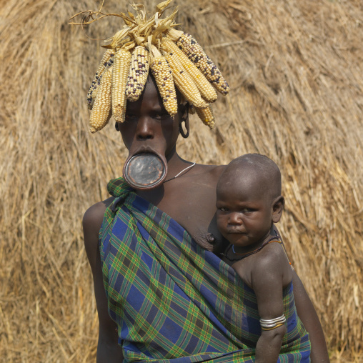 Mother Mursi Woman Wearing Corn Cob Headdress And Clay Plate Carrying Baby Ethiopia