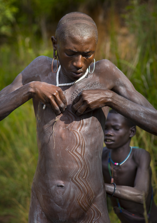 Shepherd from Suri tribe receiving help to decorate his body with camouflage paintings, Tulgit, Omo valley, Ethiopia