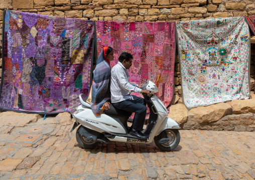 Indian couple on a scooter passing by rajasthani tapestries for sale in Jaisalmer fort, Rajasthan, Jaisalmer, India