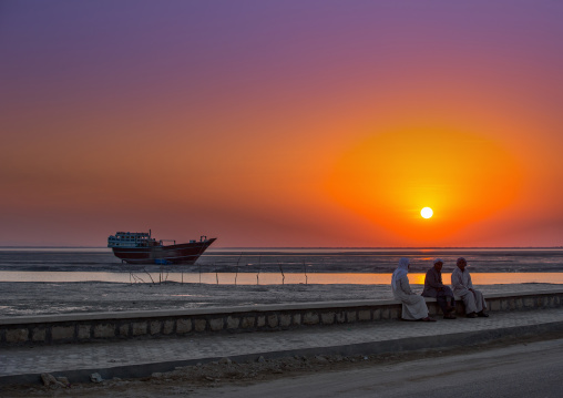 men dressed in traditional dress, sitting on a bench in front of a sunset and a dhow boat, Qeshm Island, Laft, Iran