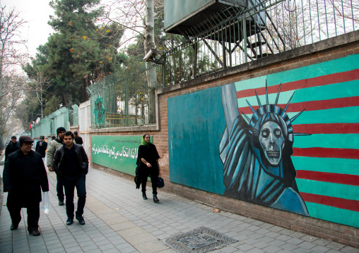 people passing in front of anti-american mural propoganda slogan depict statue liberty skeleton on the wall of the former united states embassy, Central district, Tehran, Iran