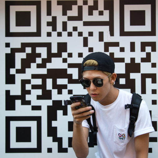 Young japanese man looking at his mobile phone in front of a giant bar code, Kanto region, Tokyo, Japan