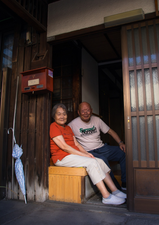 Smiling japanese couple sit in the entrance of their wooden house, Okayama Prefecture, Kurashiki, Japan