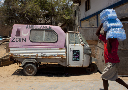 A man carrying drinkable water on his shoulders in front of an ambulance, Lamu County, Lamu, Kenya