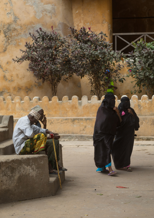 Muslim women in burqas passing in front of men sit on a bench in the street, Lamu county, Lamu town, Kenya