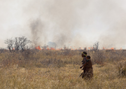 San Woman With Her Baby Passing By A Fire In The Bush, Namibia