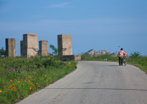 North Korean woman pushing her bicycle in front of anti tank invasion concrete blocks on roadside, Kangwon-do, Kumgang, North Korea