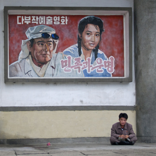 North Korean man sitting underneath a movie poster in the street, South Hamgyong Province, Hamhung, North Korea