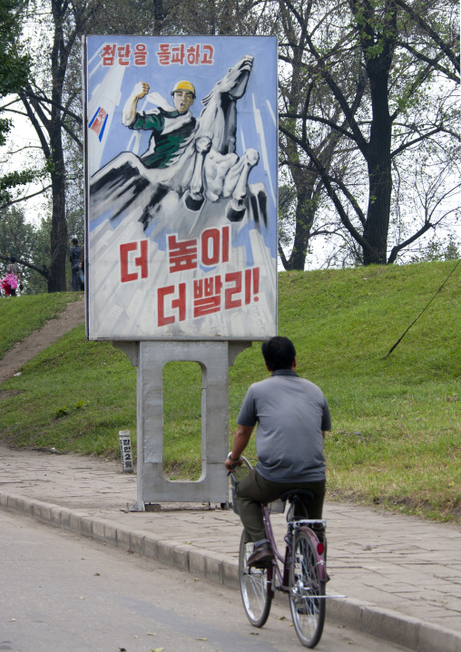 North Korean man cycling in front of a propaganda poster depicting the Chollima, Pyongan Province, Pyongyang, North Korea