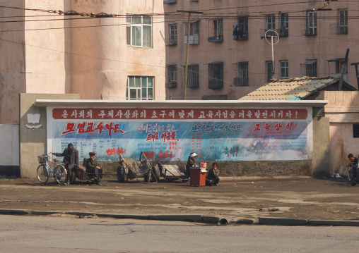 North Korean people in the street sit in front a wide propaganda billboard, South Pyongan Province, Nampo, North Korea