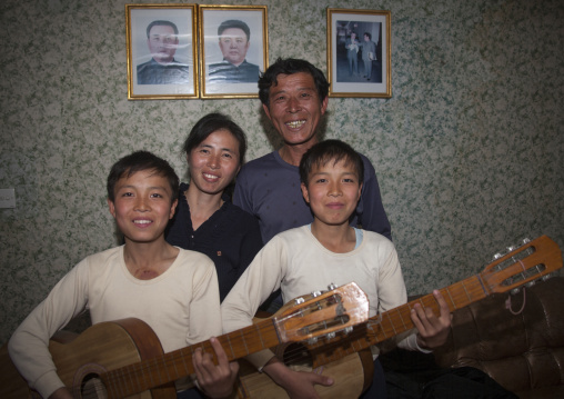 Smiling North Korean family with twins playing guitar in front of the official portraits of the Dear Leaders on the wall, North Hamgyong Province, Jung Pyong Ri, North Korea