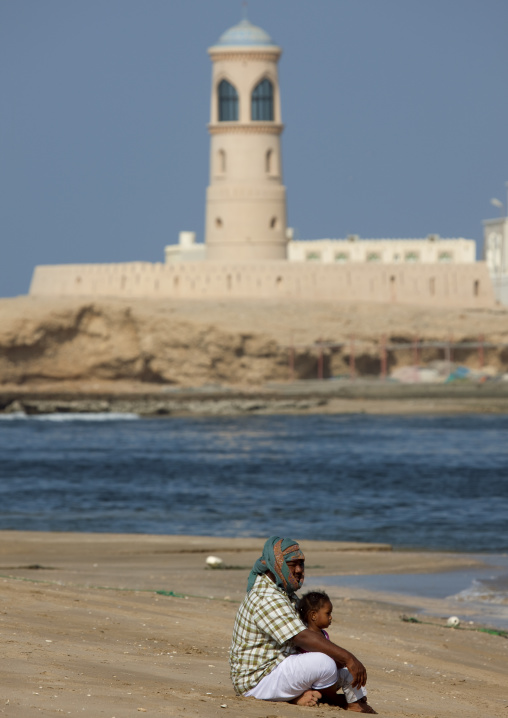 Man Sitting With His Kid Oh The Beach With The Background Of Lighthouse In Sur Port, Oman
