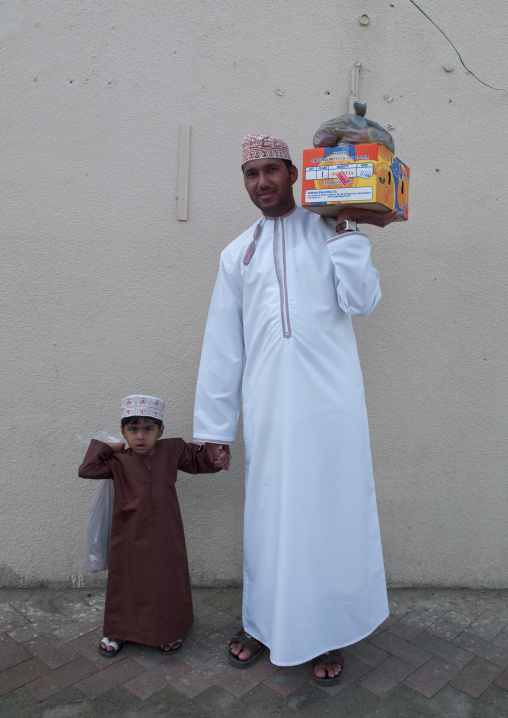 Father And Son Both Wearing Traditional Dishdasha And Carring Goods, Ibra, Oman