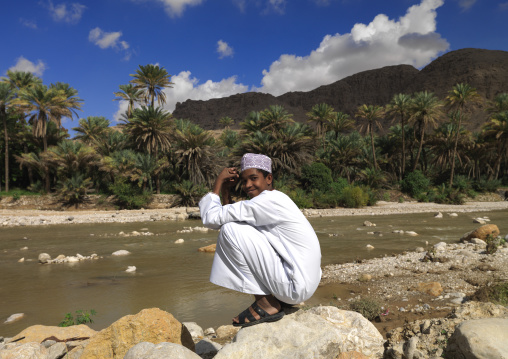 Boy Squatting On The Rock Beside The River With A Background Of Palm Trees, Wadi Bani Khalid, Oman