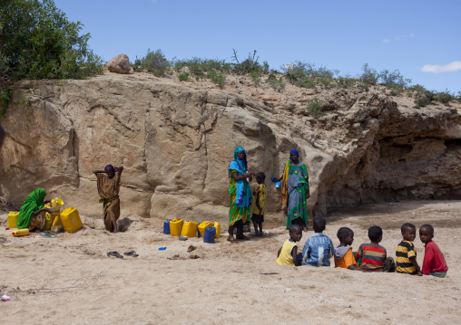 Women Taking Drinking Water From A Well Hole In The Sand And Pouring It Into Containers Children Playing Along, Lasadacwo Village, Somaliland