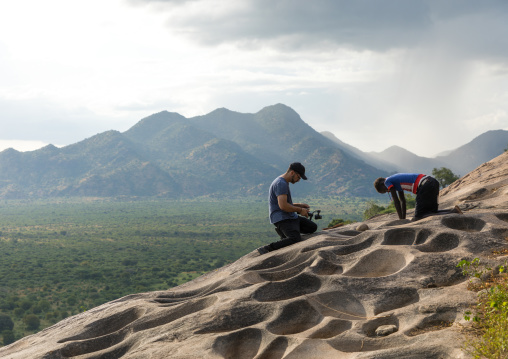 Tourist filming a Lotuko tribe woman grinding grains in a rock hole, Central Equatoria, Illeu, South Sudan
