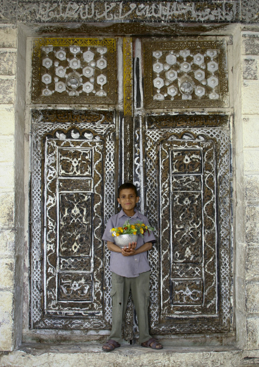 Smiling Boy Holding A Silver Ball Full Of Flowers In Front Of The Mosque Wooden Door In Ibb, Yemen