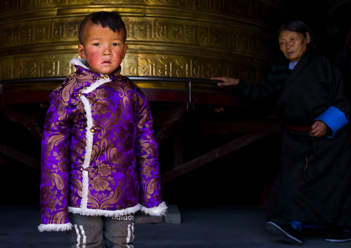 Portrait of a tibetan nomad boy with his cheeks reddened by the harsh weather in front of a prayer wheel in Rongwo monastery, Tongren County, Longwu, China