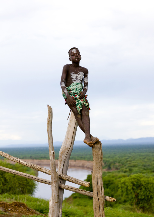 Portrait Of A Karo Tribe Kid With Body Paint Climbed On A Fence Over The Omo River, Korcho Village, Omo Valley, Ethiopia