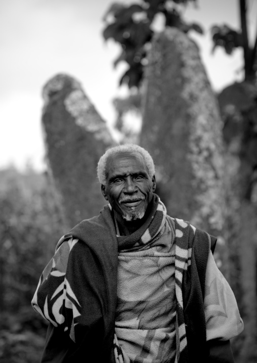 Black and white portrait of an old man in front of phallic shaped steles in gedeo area, Ethiopia