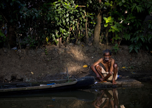 Squatting Old Man Alongside His Pirogue On A Shore On Backwaters Of Kerala, Alleppey, India