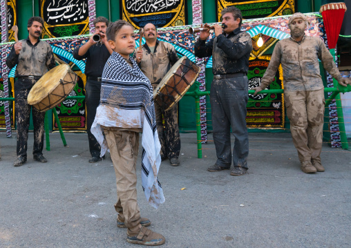Iranian shiite muslim child with mud stains on his clothes in front of musicians during the Ashura day, Lorestan Province, Khorramabad, Iran