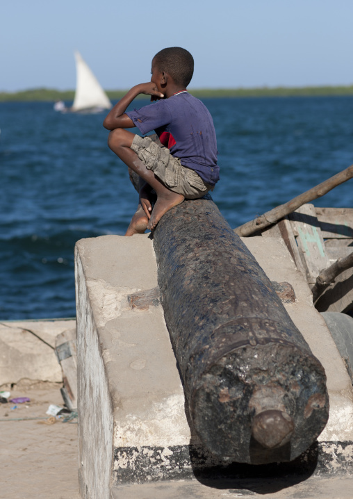 Boy sit on a cannon along a dockside, Lamu County, Lamu, Kenya