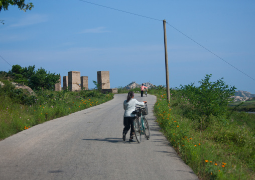 North Korean women pushing their bicycles in front of anti tank invasion concrete blocks on roadside, Kangwon-do, Kumgang, North Korea
