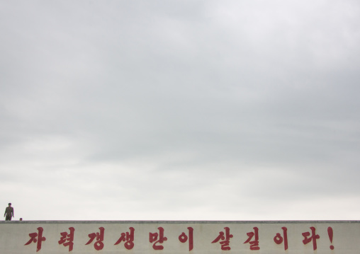 North Korean man standing above a slogan on the roof of a farm saying autonomous regeneration and recovering by one's own means are the only way to survive!, South Hamgyong Province, Hamhung,