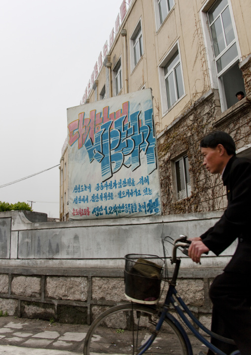 North Korean cyclist passing in front of a propaganda billboard about the great economic development
, North Hwanghae Province, Kaesong, North Korea