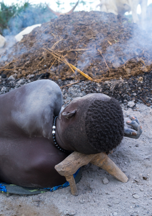Mundari tribe man resting on a wooden pillow in front of a bonfire made with dried cow dungs to repel mosquitoes, Central Equatoria, Terekeka, South Sudan